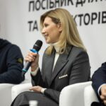перша леді тримає в руках мікрофон, сидить на стільчику на фоні банера про Альбом